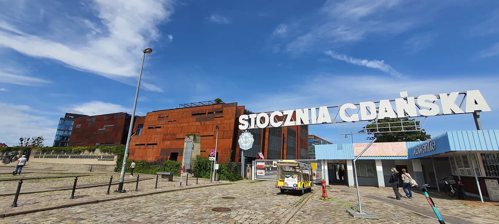 The entrance to Stocznia Gdanska, marked by a large white sign, with a modern, rust-colored building in the background under a blue sky.